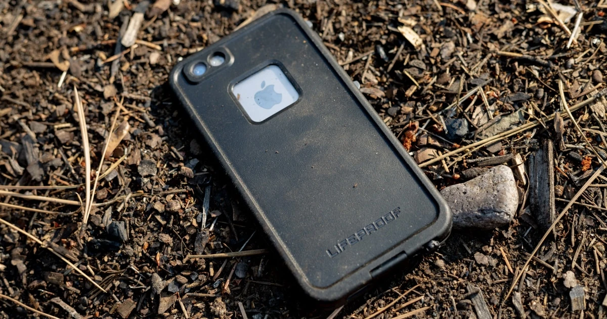 Dusty phone in a rugged case lying on dirt and wood debris.
